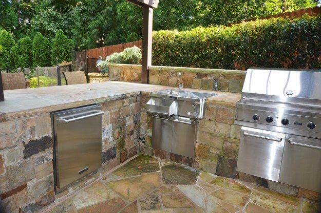 stone outdoor kitchen with steel grill, sink, and refrigerator in landscape backyard