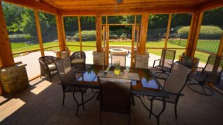 A dining table and chairs sit inside a timber screened porch overlooking a green lawn and trees.