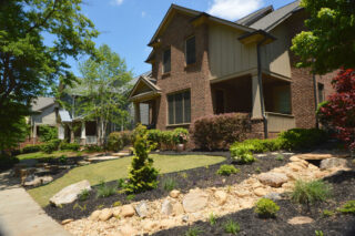 A shallow rock-lined channel runs beside a planted front yard and a house with a covered porch.