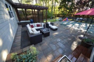 White-cushioned outdoor chairs sit on a paved patio beside a house, with a timber pergola and a red umbrella nearby.