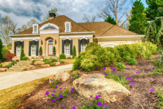 Flowering groundcover and boulders border a front lawn and pale driveway beside a house.