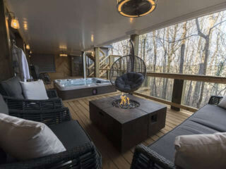 Dark woven sofas and a hanging chair surround a square table beneath a covered deck overlooking leafless trees.
