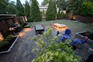 Round blue chairs and a small table sit on a paved garden patio beside a rectangular fire feature and a screened boundary.