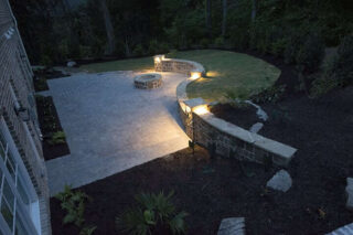 Small lights illuminate a curved stone wall along the edge of a broad paved patio beside a house.