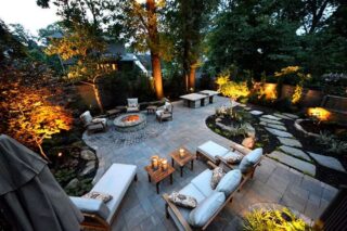 An elevated view shows cushioned outdoor seating, a fire feature, curving garden paths and trees illuminated at dusk.