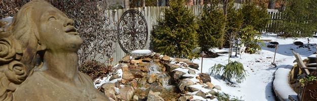 backyard stone statue with stone garden fountain and ever green trees covered in winter snow