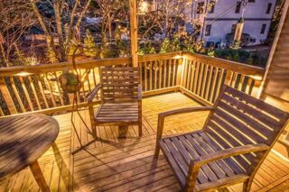 Wooden chairs and a small table sit on a timber deck with warm lighting and a railing overlooking the garden.