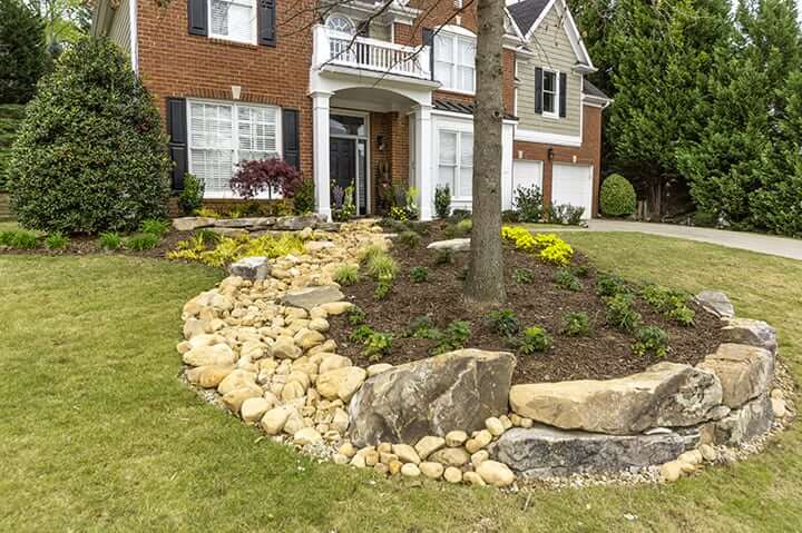 rock landscaping with trees and yellow flowers in front of brick house and green grass yard