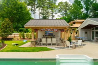 A roofed outdoor kitchen pavilion with bar stools faces a swimming pool, beside patio dining furniture and a lawn.