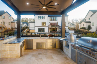 A covered outdoor kitchen includes a stainless steel grill, storage cabinets and a ceiling fan overlooking neighboring homes.