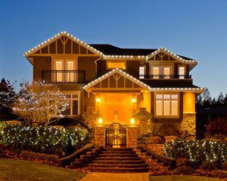 Warm lights outline the roof and entrance of a two-story house, with illuminated garden trees beneath a blue dusk sky.