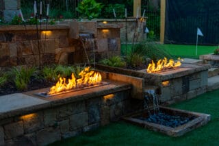 Rectangular fire features and small waterfalls set in a stone wall, with plants and turf, create a dramatic outdoor scene at dusk.