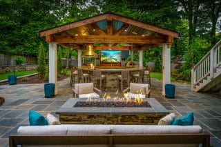 A timber pavilion shelters an outdoor dining area behind a rectangular fire feature and cushioned patio seating.