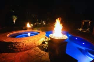 A flame rises from a raised fire bowl beside a curved blue-lit pool and round stone spa at night.