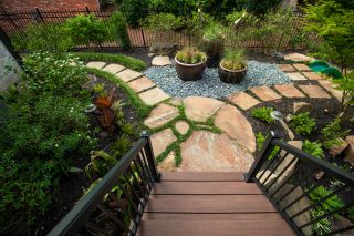 Timber steps lead down to an irregular stone patio with potted plants, gravel and dense garden planting.