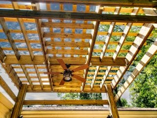 An upward view shows a grid of timber pergola beams beneath translucent roof panels with a ceiling fan below.