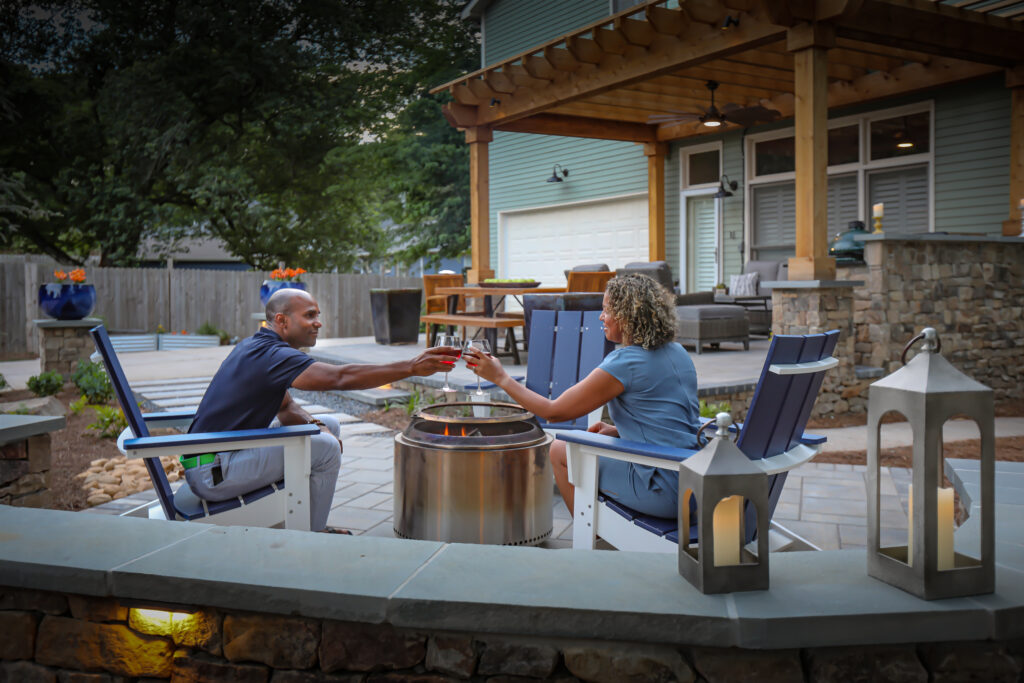A couple toasts drinks by a fire pit in a modern backyard with lanterns, stone wall, and covered patio at dusk.