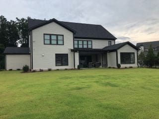 Modern two-story house with light siding, large windows, covered patio with outdoor furniture, and a spacious green lawn in front.
