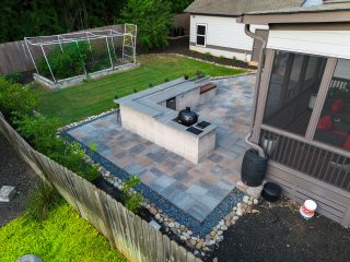 An overhead view shows an outdoor kitchen island on a gray paved patio beside lawn, a screened porch and a fenced garden.