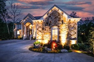 Stone house at dusk with warm lights, arched doorway, large window, tidy landscaping, driveway, and a colorful sky in the background.