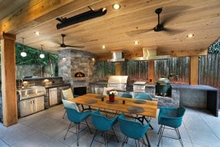 An Atlanta-style covered outdoor kitchen with stone grill, pizza oven, wood ceiling, blue chairs at a wooden table, and lush greenery outside.