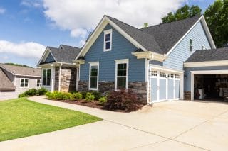 A modern blue house with stone accents, white trim, and large garage doors sits behind neat shrubs and a manicured lawn on a sunny day.