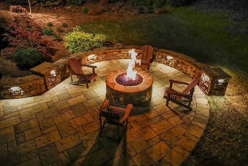 Chairs surround a lit round fire pit on a curved paved patio bordered by low stone walls at night.