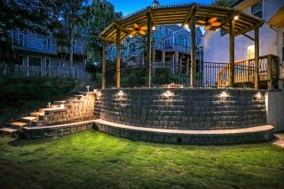 A timber pergola stands above a curved stone retaining wall with illuminated steps leading up from the lawn at dusk.