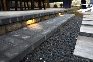 Outdoor stone steps with built-in yellow lights lead from a patio with a table and chairs down to a gravel area with large stepping stones.