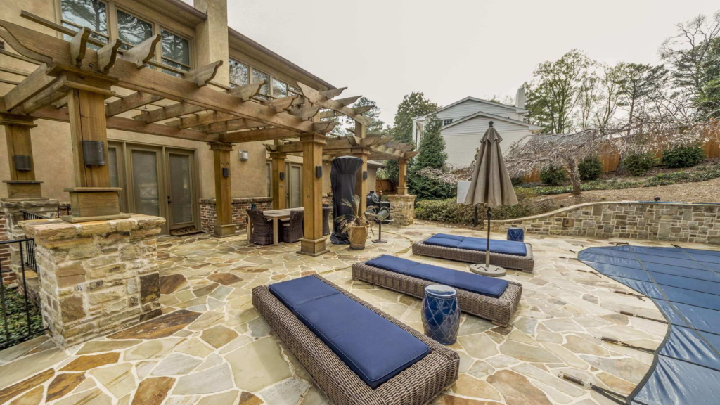 Blue-cushioned lounge chairs line a stone pool terrace beside a house and timber pergola.