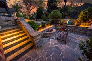Stone patio at sunset with lit steps, a fire pit, wooden benches, lush plants, and large blue pots creates a cozy outdoor setting.