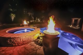 Luxurious backyard at night with a lit fire bowl, glowing hot tub, blue-lit pool, elegant stonework, and landscaped surroundings.