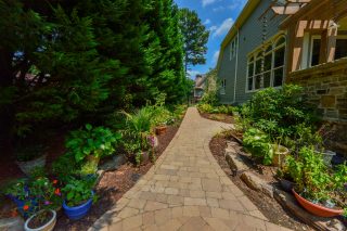 A stone path curves through a lush garden beside a house, lined with potted plants and greenery beneath a sunny blue sky.