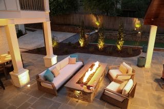 Sofas and chairs surround a rectangular fire feature on a covered patio, with illuminated planting along the fence beyond.