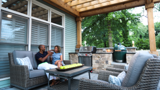 Two people sit on an outdoor sofa beneath a timber roof beside a grill, counter and matching woven chairs.