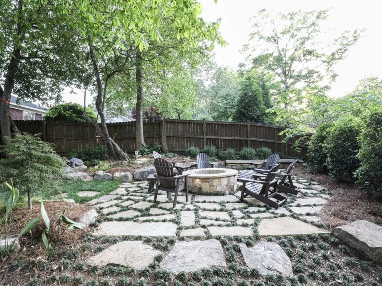 A backyard patio features stone pavers, a circular fire pit, Adirondack chairs, and is bordered by trees, plants, and a wooden fence.