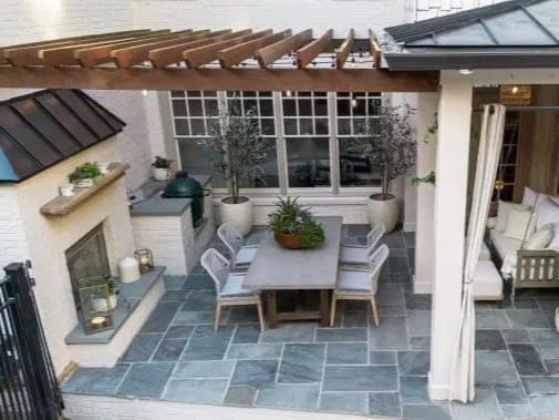 A modern patio with a pergola, dining table and chairs, potted plants, fireplace, slate tile flooring, and cushioned corner seating.