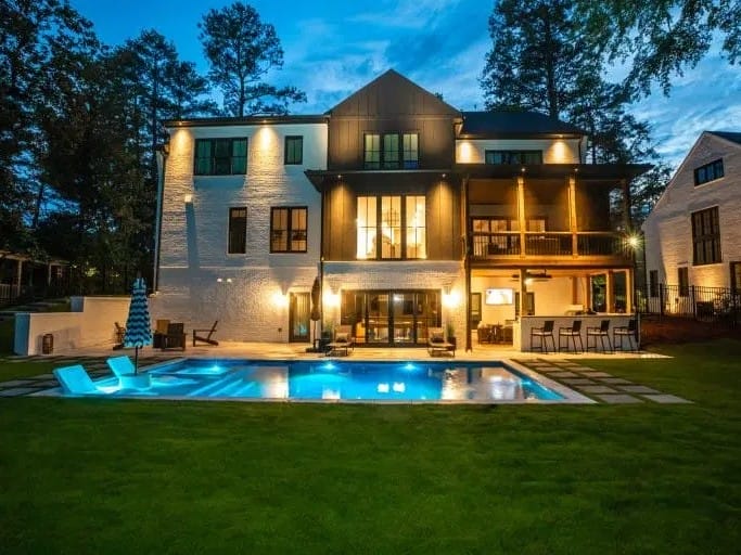 A large modern house at dusk features bright outdoor lighting, a balcony, and a pool with loungers on a grassy yard.