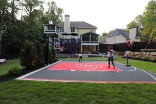 A person stands on a red and gray backyard athletic court with a basketball hoop, surrounded by lawn and neighboring homes.