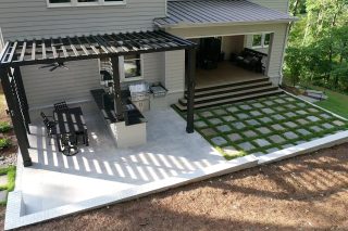 An elevated view shows a dark pergola over an outdoor kitchen and dining space beside a checkerboard pattern of paving and grass.