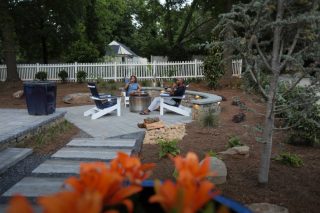 Lounge chairs surround a fire feature on a garden patio, with people seated nearby and orange flowers in the foreground.