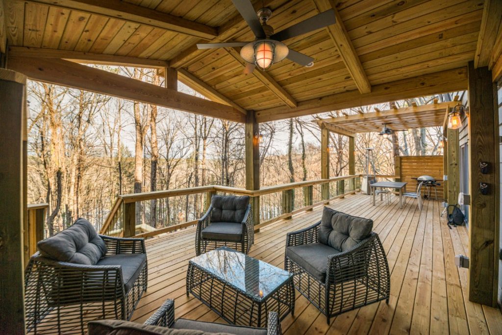 A covered wooden deck with black wicker furniture, a glass coffee table, ceiling fan, and nearby grill area surrounded by leafless trees.