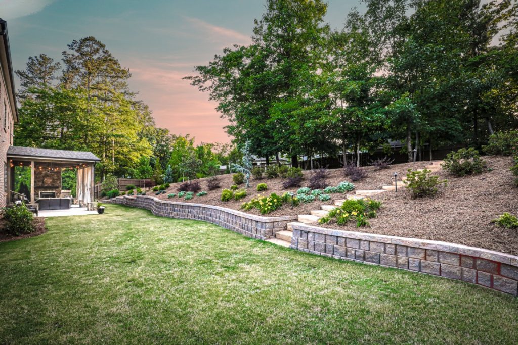 A landscaped backyard features tiered stone walls, mulched beds with shrubs and trees, a covered patio on the left, steps up the slope, and a pink-blue sky.