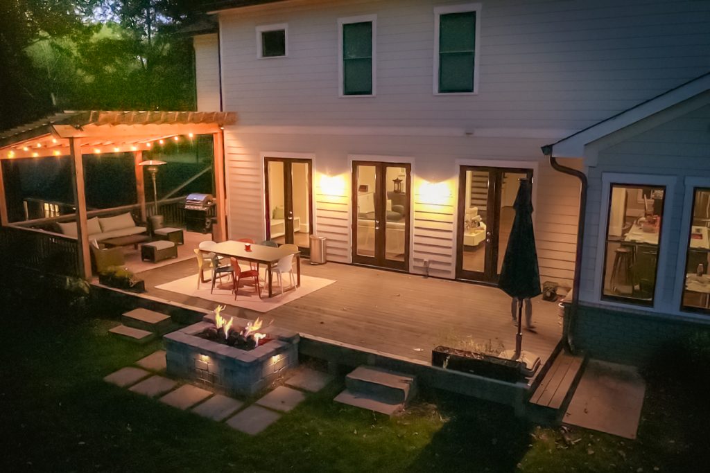 Backyard deck at dusk with a pergola, string lights, dining table, fire pit, outdoor seating, grill, and a white house with lit glass doors.