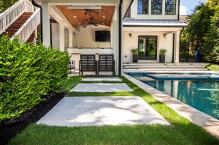 Wide rectangular paving slabs separated by grass lead beside a swimming pool toward a covered patio and house.
