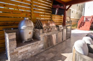 A stone-faced outdoor kitchen with metal cooking appliances and cabinet doors sits beneath a timber roof and slatted screen.