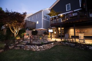 A cozy backyard at dusk features a firepit, Adirondack chairs, patio lights, stone landscaping, and a dark gray house with glowing windows.