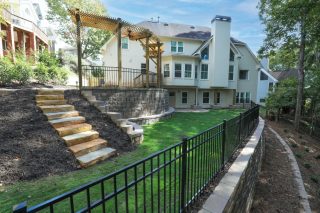 Backyard of a large house featuring stone steps, a metal fence, a wooden pergola on a raised patio, and landscaped gardens with grass and trees.