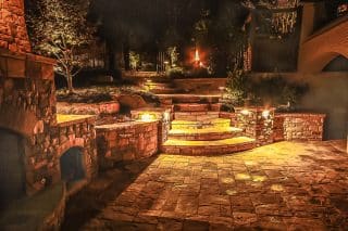 A warmly lit stone patio with curved steps, stone walls, and lush plants glows at night as small lights highlight the path and garden.