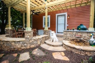 A fluffy white dog sits on a stone patio with a table, chairs, grill, and landscaping in front of a red house with a white door.